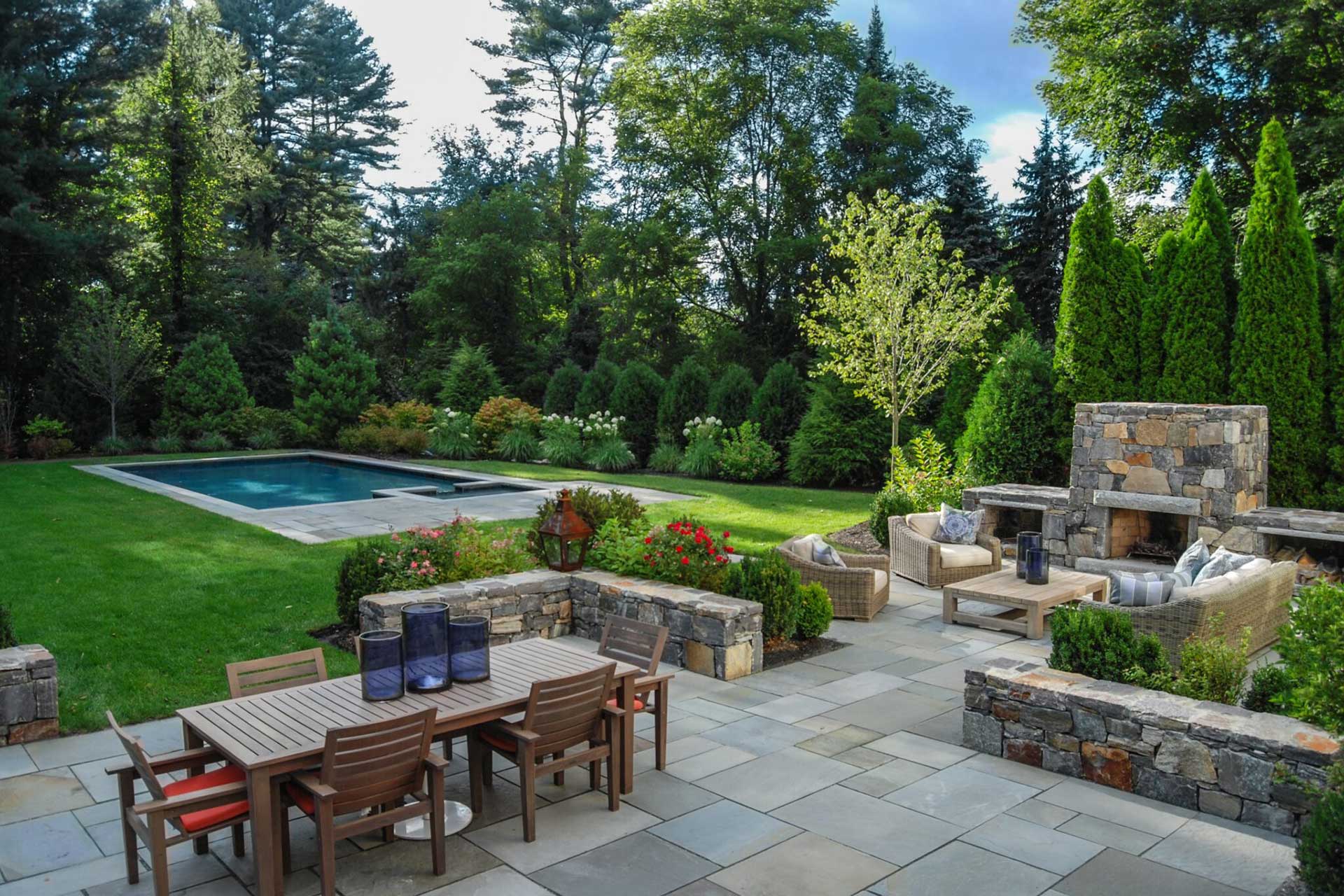 After photo of renovated Weston, MA backyard with bluestone patio, pool, stone fireplace, and evergreen screening.